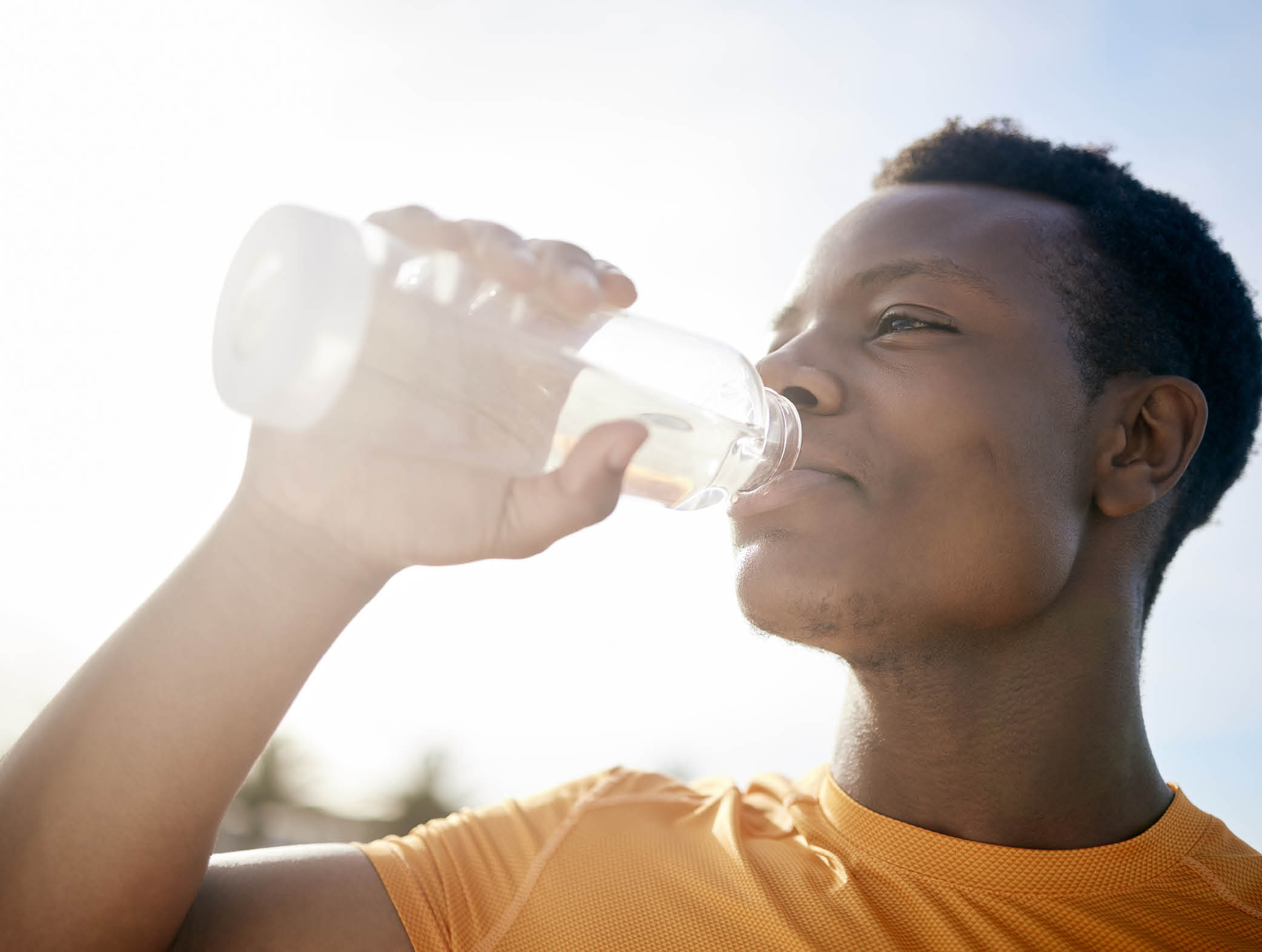 A jogger drinking a bottle of water in the sunlight. An active, healthy African American man quenching his thirst after exercising