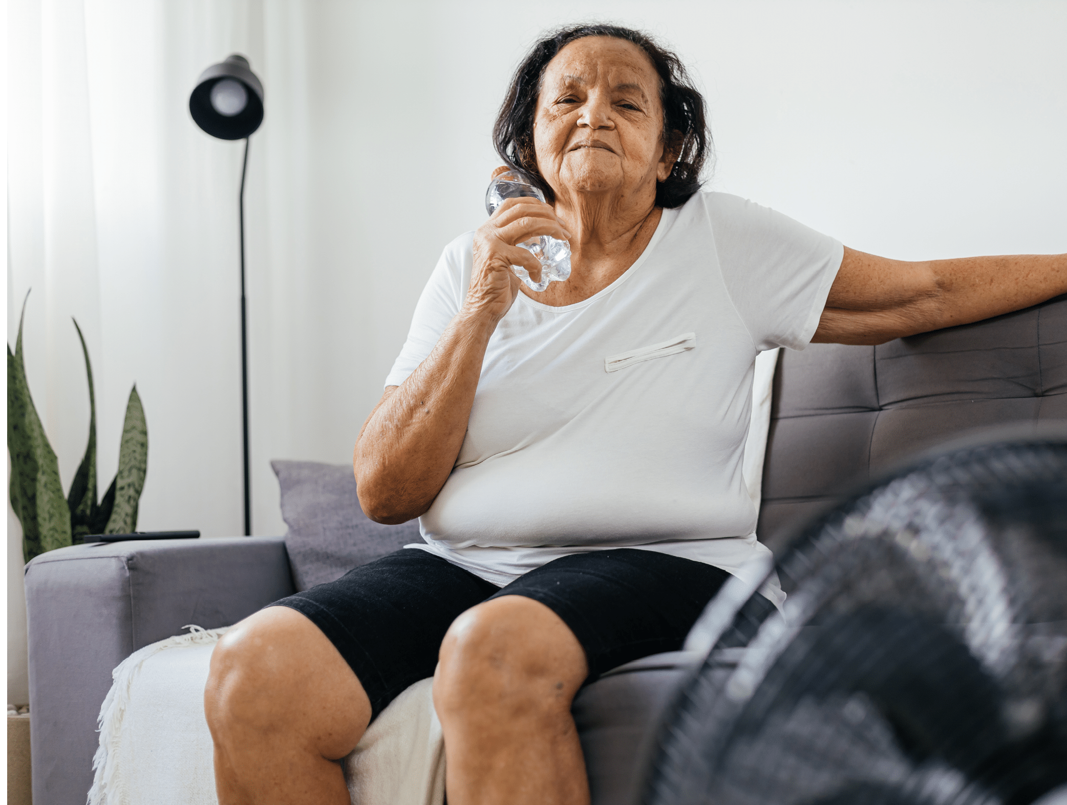 Elderly woman sitting on sofa in living room cooling off with floor fan trying to relieve heat of hot summer weather. Elderly woman cooling her face with a bottle of cold water.